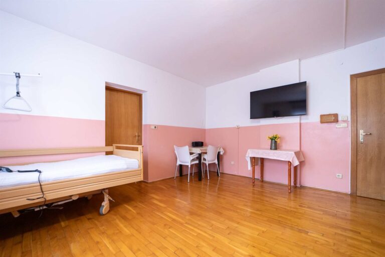 velika spavaća soba u domu za starije osobe sa stolom, televizorm, i krevetom za starije i nemoćne osobe